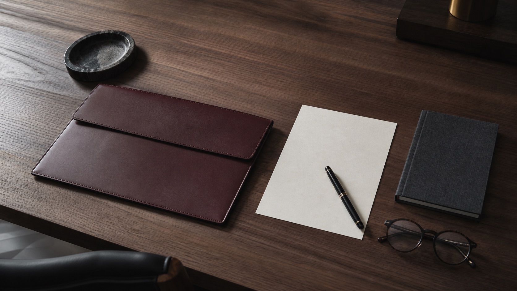 Tavolo da lavoro in legno scuro con taccuino, penna, occhiali e portadocumenti sobri disposti con ordine.